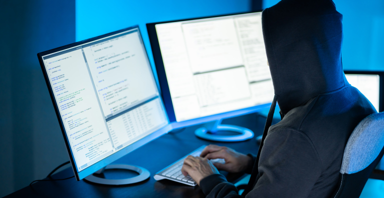 A person with a hood over their head looks at two computer screens with numbers and codes displayed.