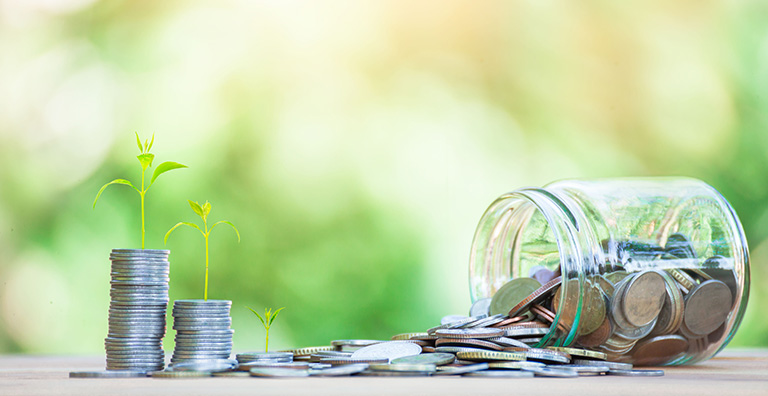 A tipped over jar of quarters next to a stack of quarters surrounded by green plants.